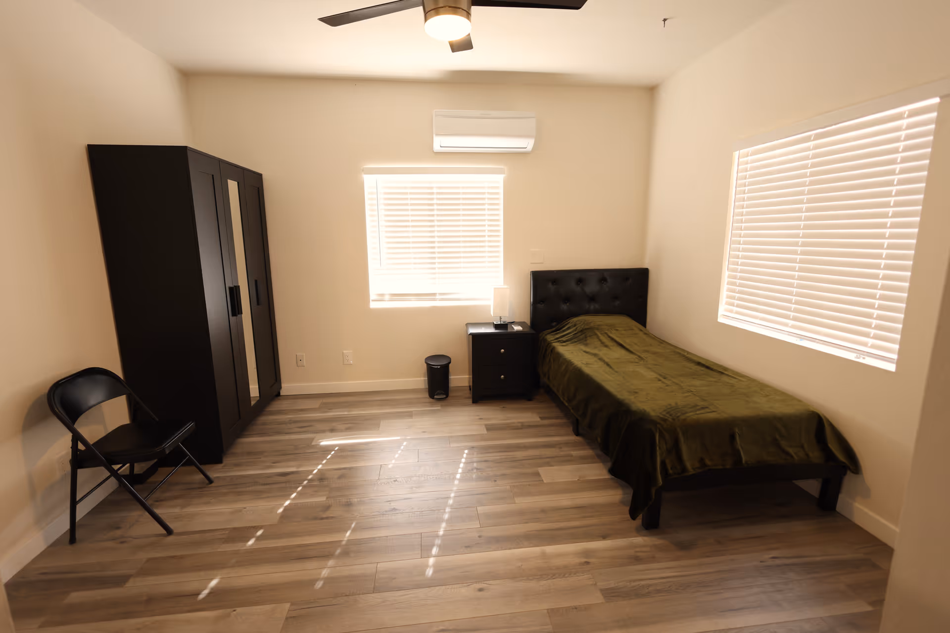 Simple furnished bedroom with a single bed, nightstand, wardrobe, folding chair, and wood-look flooring.