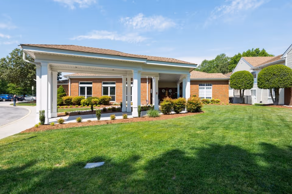 Exterior view of The Gardens of Virginia Beach facility showing a covered entrance with white columns, a brick building, well-maintained green lawn, trimmed bushes, and trees under a partly cloudy blue sky.