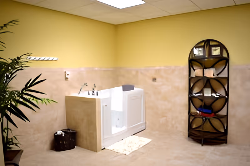 A spacious bathroom with beige tiled walls and floor, featuring a white walk-in bathtub with grab bars. There is a small dark basket on the floor next to the tub, a tall metal shelf with various items on the right, and a green plant partially visible on the left. The walls are painted yellow above the tile, and the ceiling has white tiles with recessed lighting.