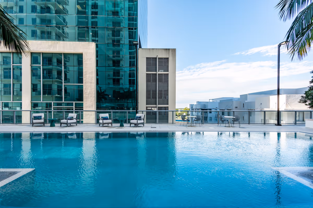 Outdoor swimming pool with clear blue water, surrounded by a deck with lounge chairs and small tables. Modern buildings with glass windows and balconies are visible in the background under a clear blue sky. Palm tree leaves frame the image on the left and right edges.