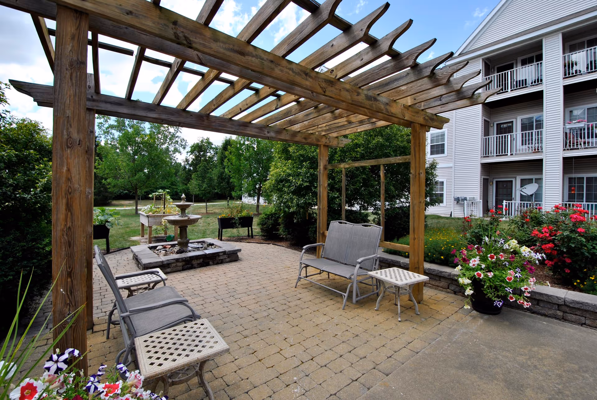 Outdoor patio area with a wooden pergola, metal chairs, a small table, a stone fountain, and various flower pots. In the background, there is a multi-story residential building with balconies and greenery surrounding the patio.