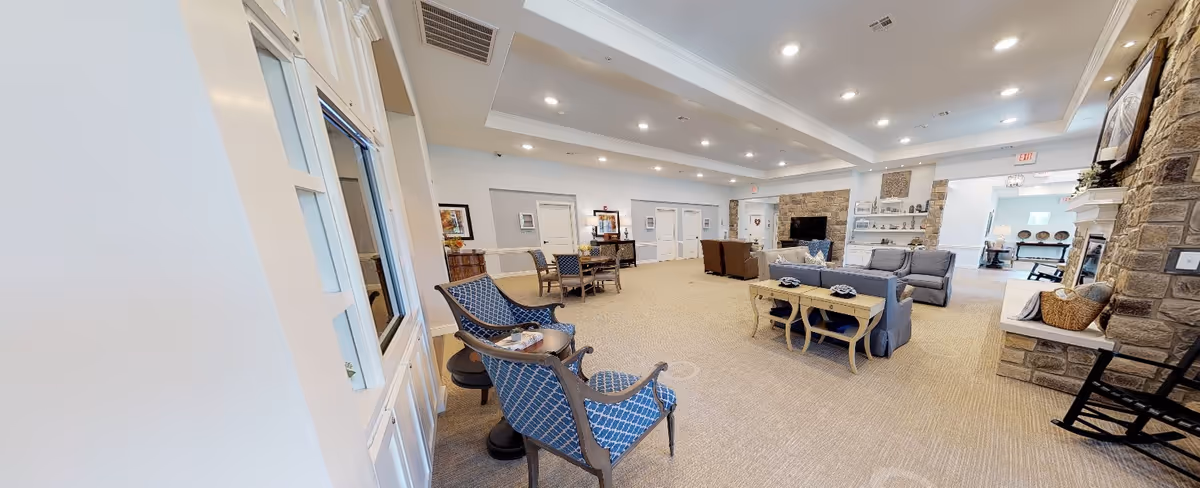 Spacious and well-lit common area in a senior living facility featuring multiple seating arrangements including blue patterned armchairs, sofas, and tables. The room has beige carpeting, recessed ceiling lights, stone accent walls, a fireplace, and a television. There are decorative items on shelves and a door leading to another room in the background.