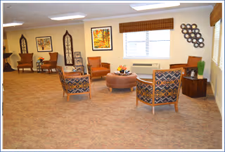 A spacious living room area with patterned armchairs arranged around a round ottoman. The room has beige carpet, cream-colored walls, and several pieces of framed artwork and decorative items on the walls. A window with blinds and a valance lets in natural light.