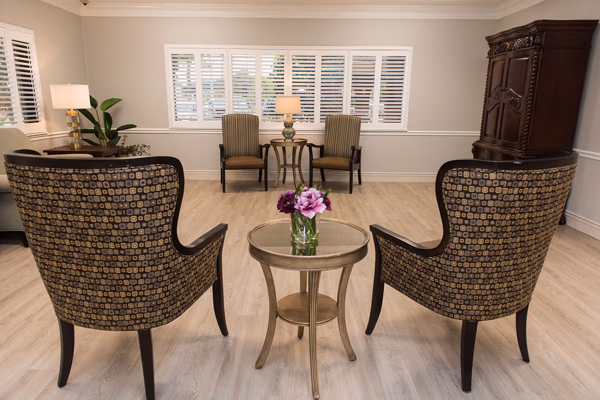 A cozy sitting area with two patterned armchairs facing a small round table with a vase of purple flowers. In the background, there are two striped armchairs with a small table and lamp between them, a large wooden cabinet, and windows with white shutters letting in natural light.