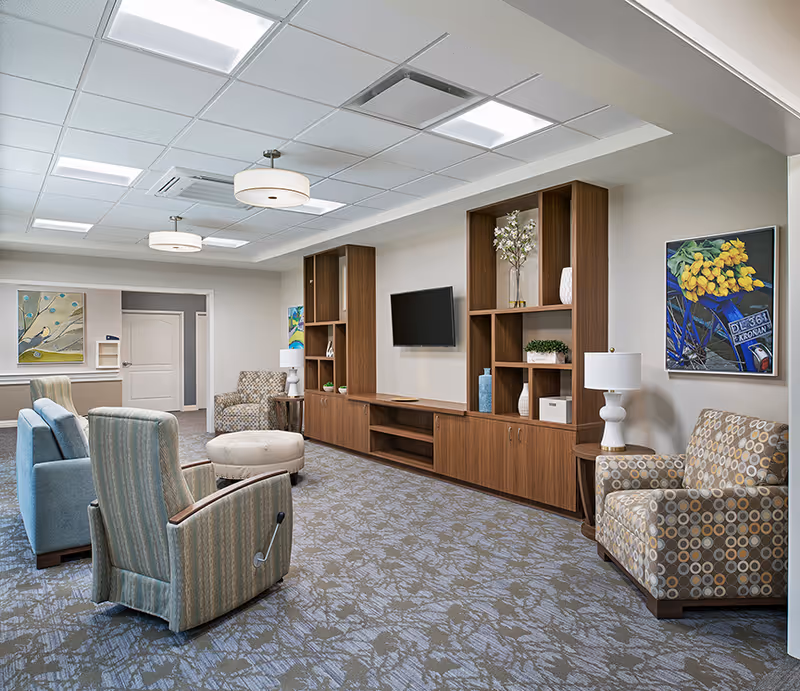 A bright and spacious living room area in a senior living facility featuring comfortable upholstered chairs, a round ottoman, a wooden entertainment center with shelves and cabinets, a wall-mounted flat-screen TV, decorative vases and plants, two table lamps, and colorful artwork on the walls.