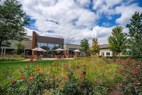 Outdoor garden area with blooming red flowers and green shrubs in the foreground, patio tables with umbrellas, and a modern single-story building under a partly cloudy sky in the background.