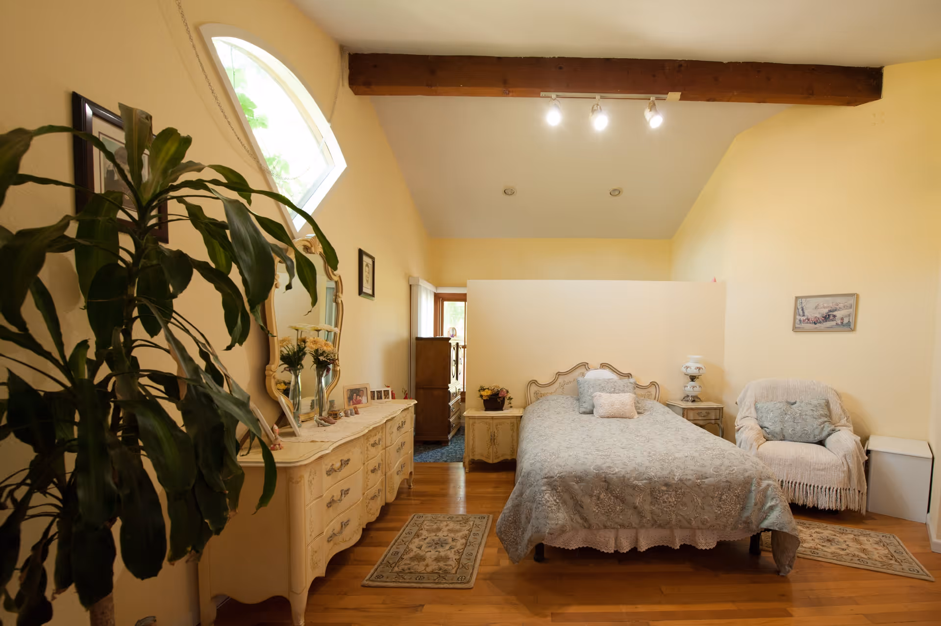 A cozy bedroom with a wooden floor, a bed with a decorative headboard and a light blue patterned bedspread, a cushioned armchair with a throw blanket, a vintage dresser with a mirror and various decorative items, a large green plant in the foreground, and soft yellow walls with a wooden beam ceiling.