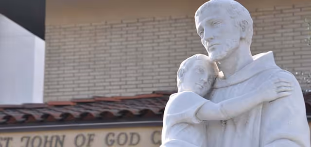 A white statue depicting a man holding a child, with a building wall and a sign partially visible in the background that reads 'ST JOHN OF GOD'.