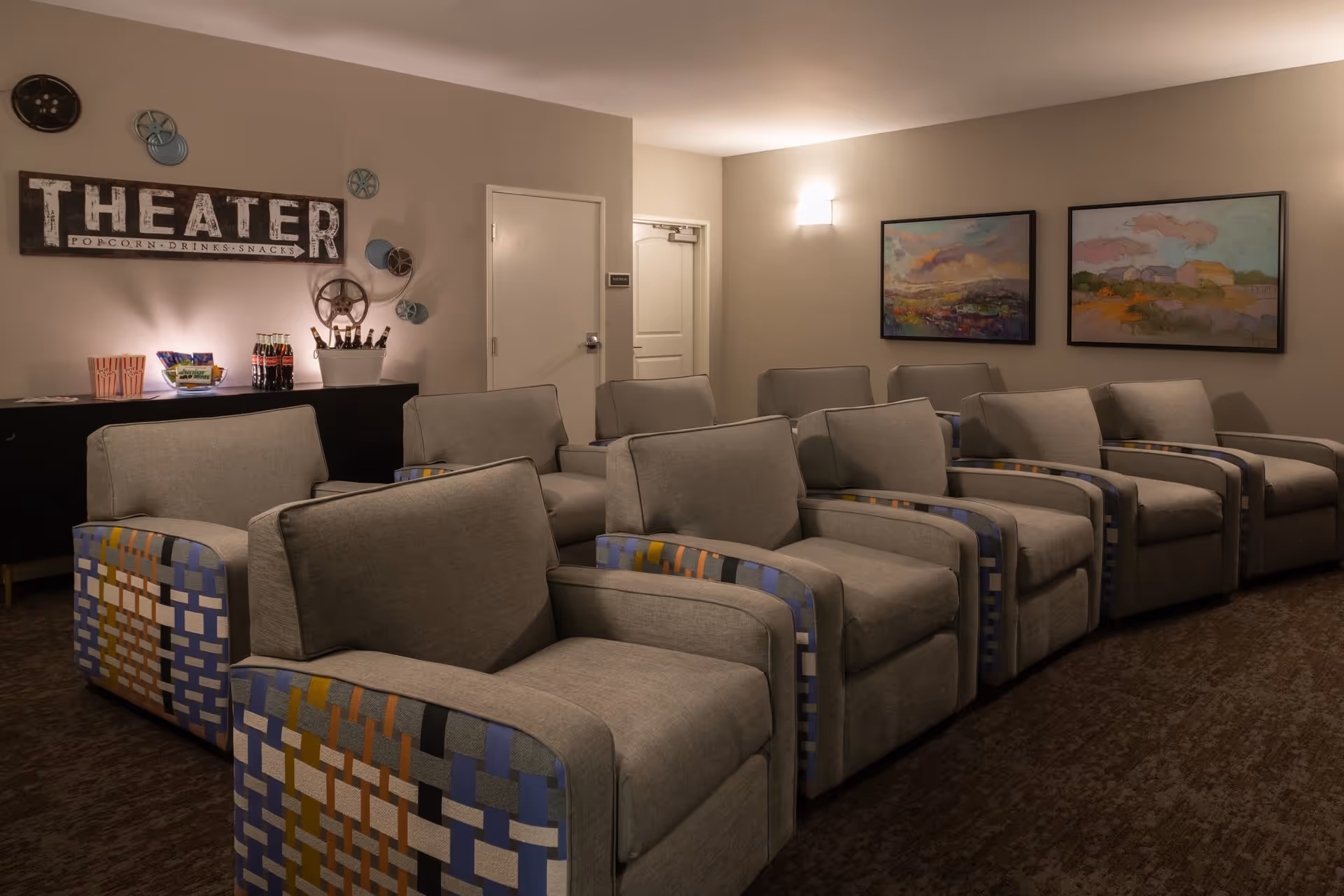 A small theater room with two rows of comfortable gray armchairs featuring patterned sides. On the left wall, there is a decorative sign that reads 'THEATER POPCORN DRINKS SNACKS' along with several film reels mounted around it. Below the sign, a black cabinet holds popcorn containers, snacks, and bottled drinks. The room has beige walls with two colorful abstract paintings on the right wall and soft lighting.
