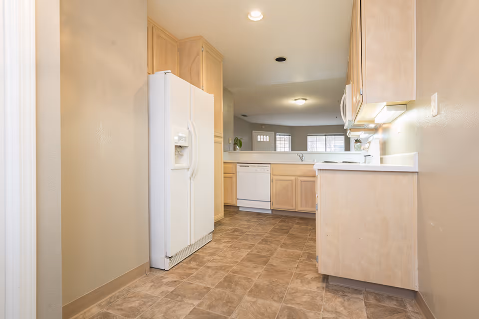 A bright kitchen with light wood cabinets, a white refrigerator with an ice dispenser, a white dishwasher, and a white stove. The kitchen has tiled flooring and beige walls. In the background, there is a living area with windows and a ceiling light.