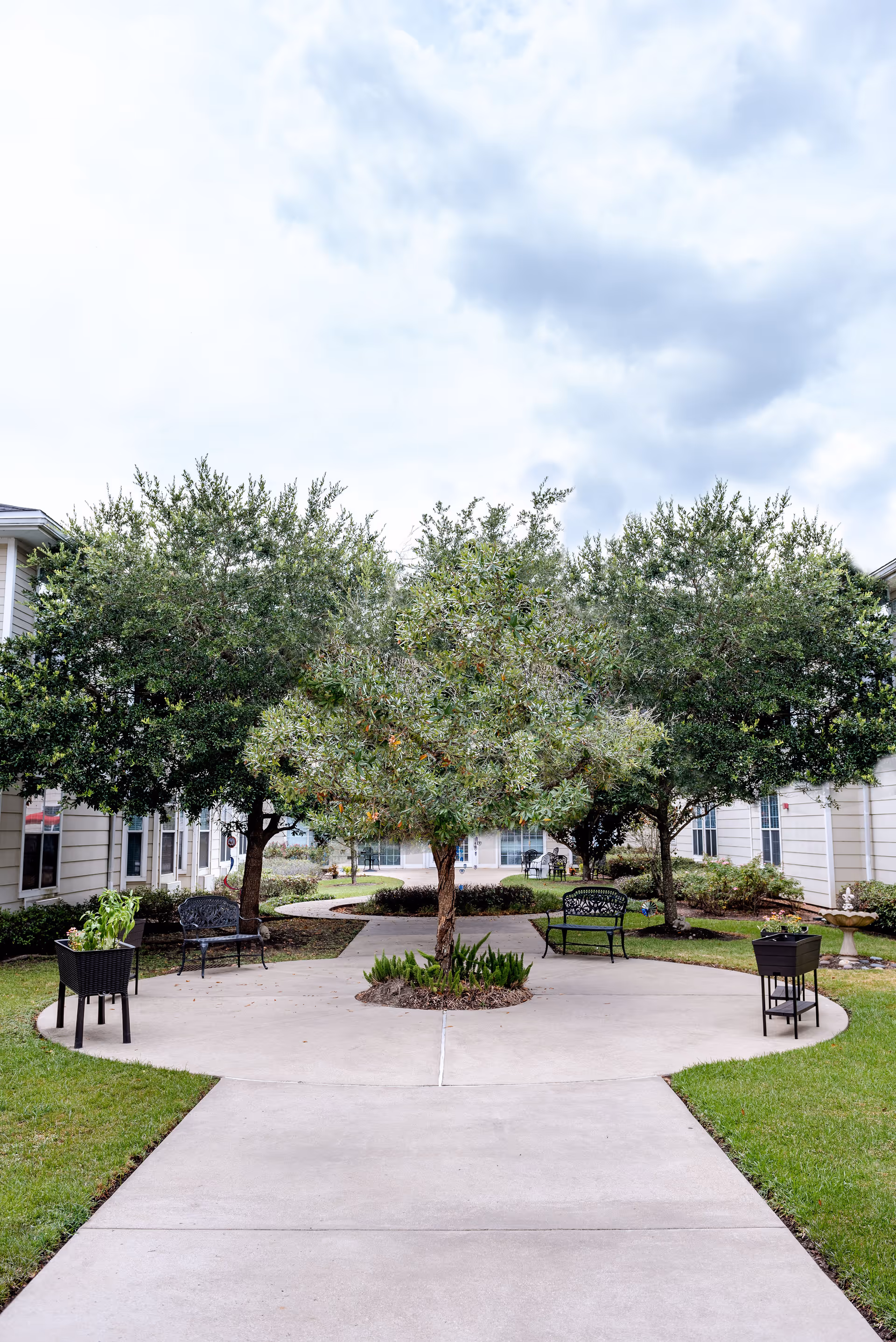 Outdoor courtyard area at The Reserve at Pearland Assisted Living and Memory Care featuring a concrete pathway surrounded by green grass, several trees, black metal benches, and planters. The courtyard is flanked by light-colored buildings under a partly cloudy sky.