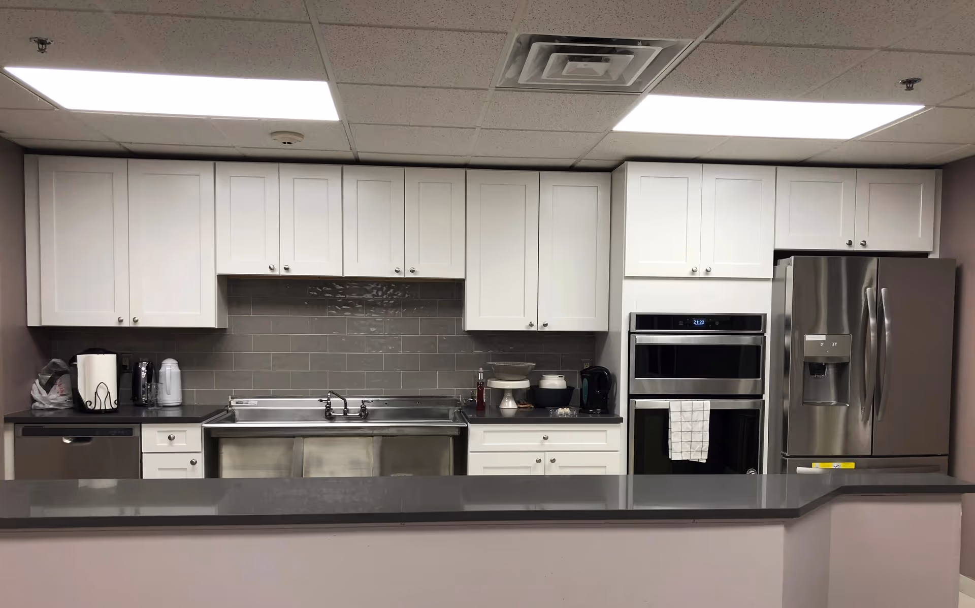 A modern kitchen with white cabinets, a stainless steel double sink, a stainless steel refrigerator with water dispenser, a built-in oven, and a dishwasher. The backsplash is made of gray subway tiles, and the countertop is a dark gray solid surface. Various kitchen items including a paper towel holder, coffee maker, and bowls are placed on the counter.