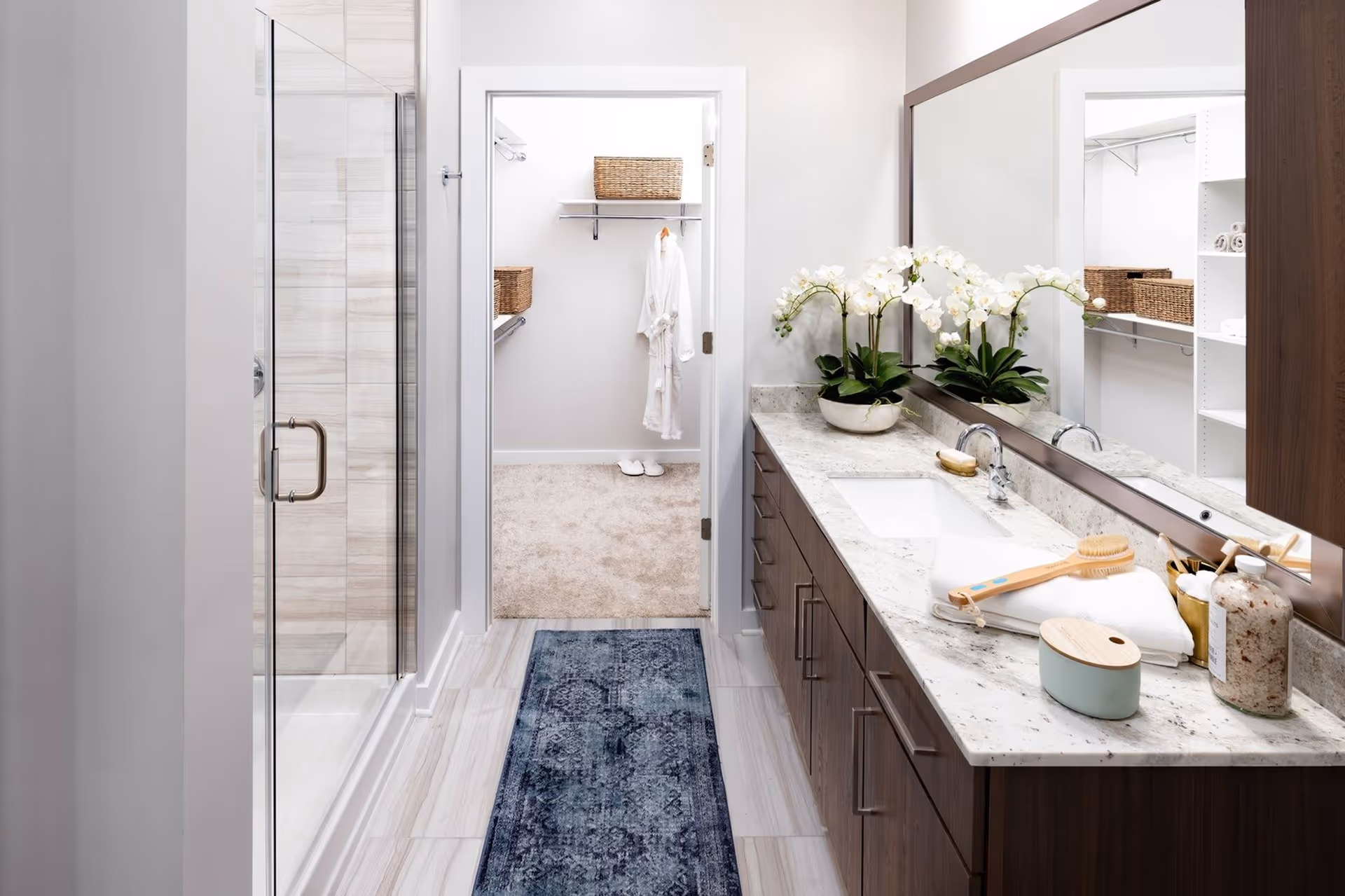 Modern bathroom with a long marble-topped vanity and large mirror, glass-enclosed shower, and a doorway leading to a walk-in closet.