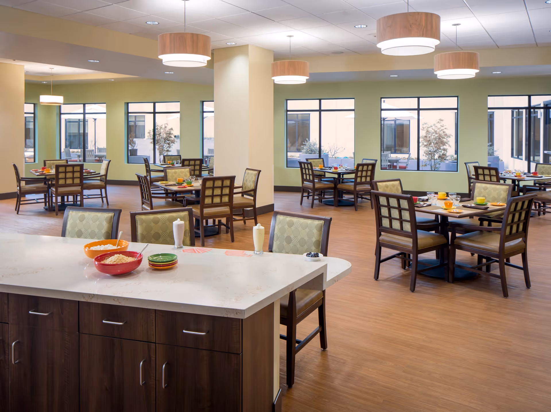A spacious dining room with multiple tables and chairs arranged neatly. The tables are set with colorful plates, bowls, and glasses. The room has large windows letting in natural light, light green walls, wooden flooring, and modern ceiling lights. In the foreground, there is a kitchen island with bowls and plates on it.