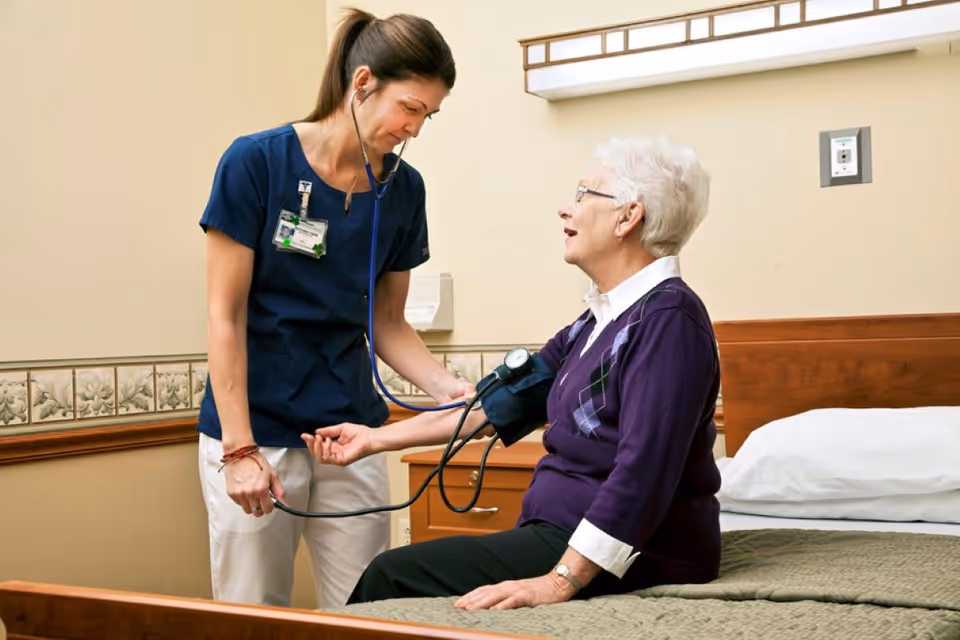 A healthcare professional in navy scrubs is measuring the blood pressure of an elderly woman with white hair who is sitting on a bed in a room with beige walls and floral wallpaper border.