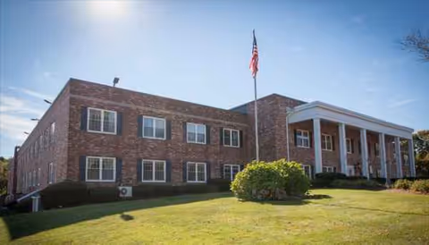 Brick senior living building with front steps, columns, flag, and well-kept lawn.