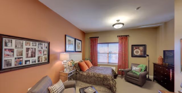 A cozy bedroom in a senior living community featuring a bed with patterned bedding and orange pillows, two armchairs with cushions, a window with red curtains, framed pictures on the walls, a bedside table with a lamp and flowers, and a dresser with a television on top.