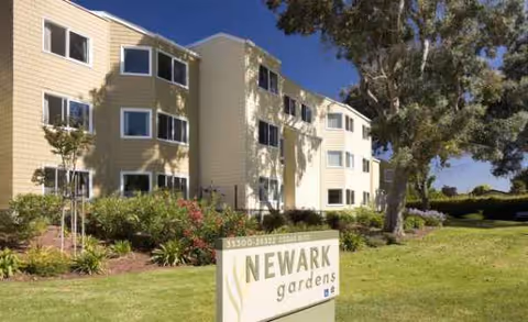 Exterior view of Newark Gardens senior living facility with sign, greenery, and trees.