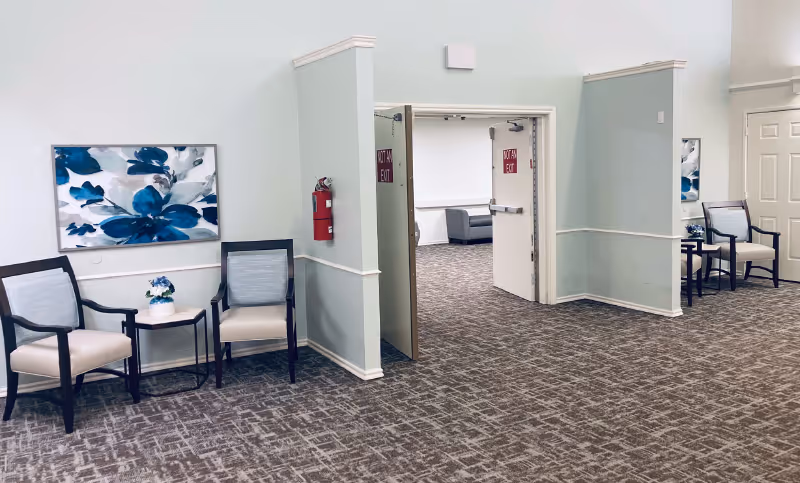 A spacious interior hallway area with light-colored walls and carpeted floor. There are two seating areas with two chairs and a small table holding a flower arrangement on each side of the hallway. Blue and white floral paintings hang on the walls above the chairs. A fire extinguisher is mounted on the wall near an open door marked with 'NOT AN EXIT' signs, leading to another room with additional seating visible.