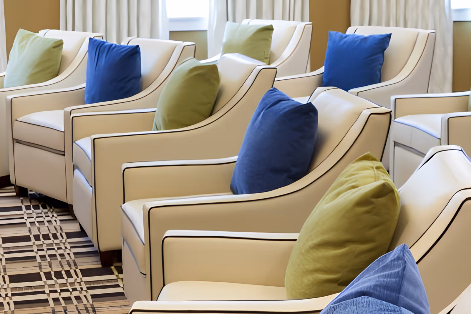 A row of cream-colored armchairs with black piping, each adorned with alternating blue and green throw pillows, arranged in a well-lit room with windows and light-colored curtains in the background.