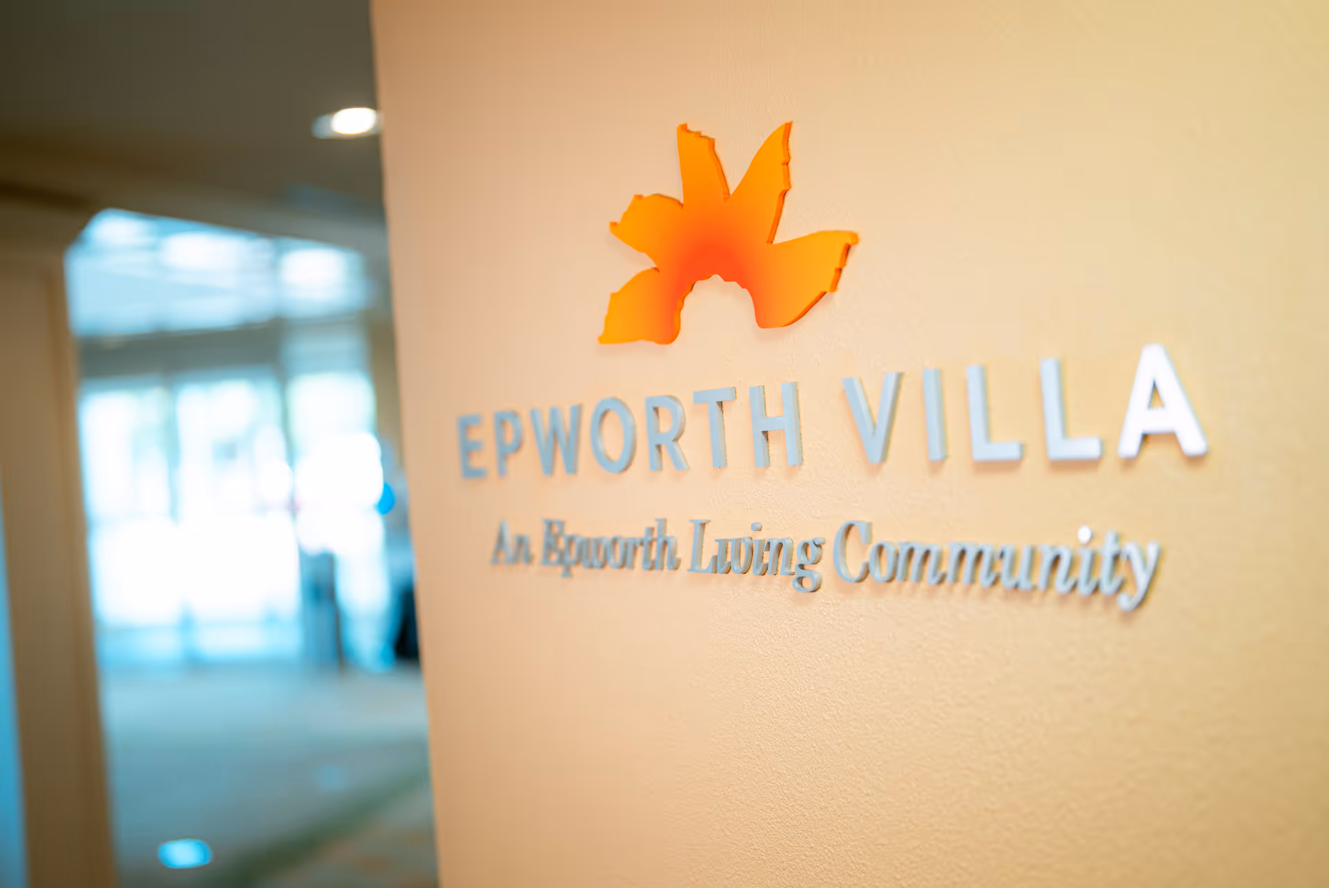 Close-up view of a wall inside a facility with the logo and text 'EPWORTH VILLA An Epworth Living Community' mounted on it, with a blurred background showing part of the interior space.