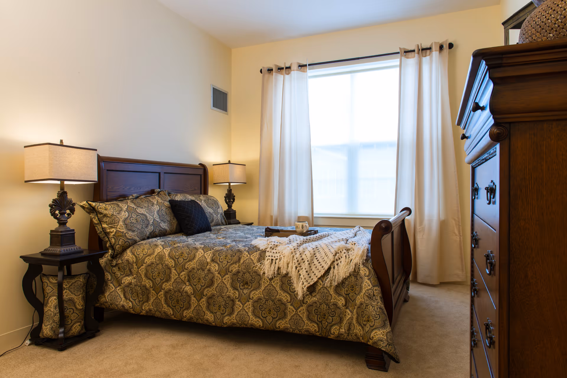 A cozy bedroom with a wooden bed frame and patterned bedding. Two matching bedside tables each hold a lamp with a beige shade. A large window with white curtains allows natural light to fill the room. A wooden dresser is visible on the right side of the image.