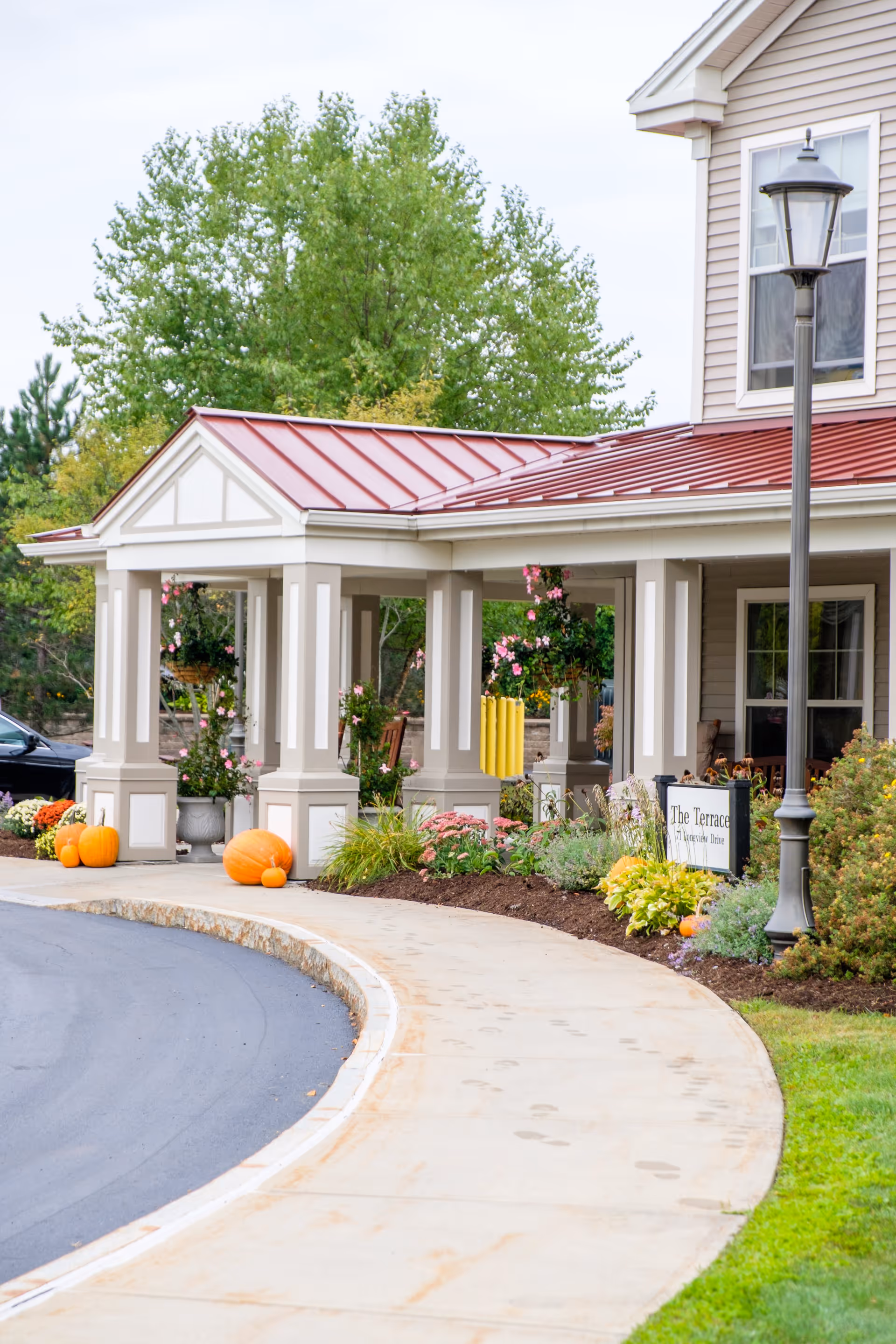 Entrance of The Terrace at The Glen facility with a covered porch supported by columns, decorated with pumpkins and hanging flower baskets. There is a curved sidewalk leading to the entrance, a lamppost, and greenery surrounding the area.