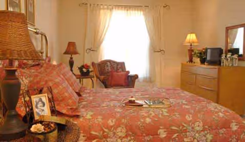 A cozy bedroom with a floral patterned bedspread on a bed, a wicker chair with a cushion near a window with sheer curtains, a dresser with a lamp, a small TV, and decorative items including framed photos and flowers.