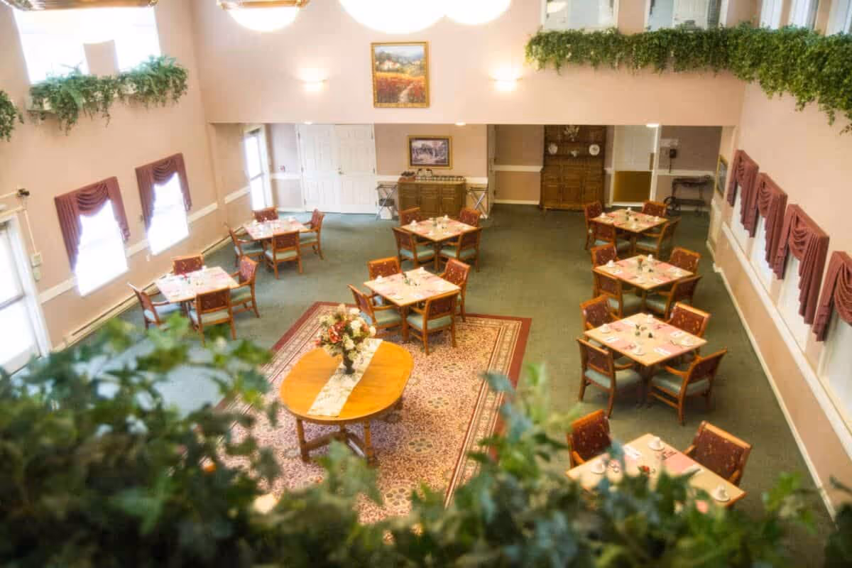 View of a dining room with several square tables each set with cups and napkins, surrounded by wooden chairs with patterned cushions. The room has green carpet, pink walls, windows with maroon curtains, and plants along the upper ledge. A round table with a floral centerpiece is in the center on a patterned rug.