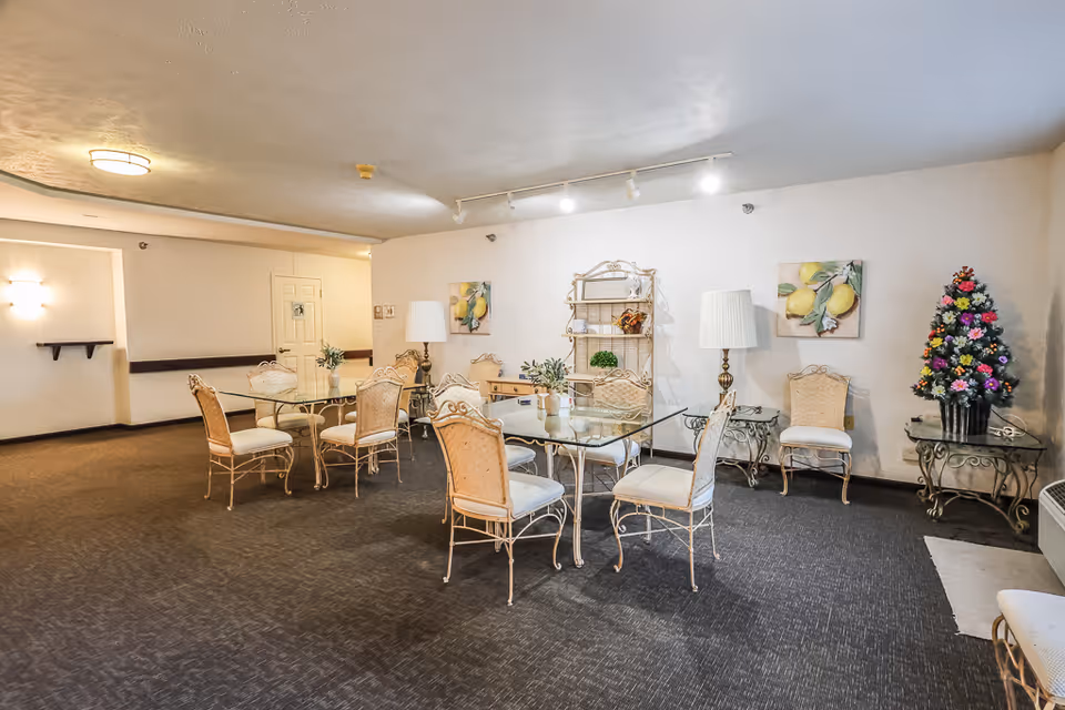 Communal dining room with glass-top tables, ornate chairs, lamps, artwork, and a small decorated Christmas tree.
