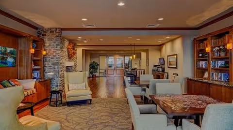 Communal lounge with comfortable chairs and tables, bookshelves, and a stone fireplace in a senior living facility.
