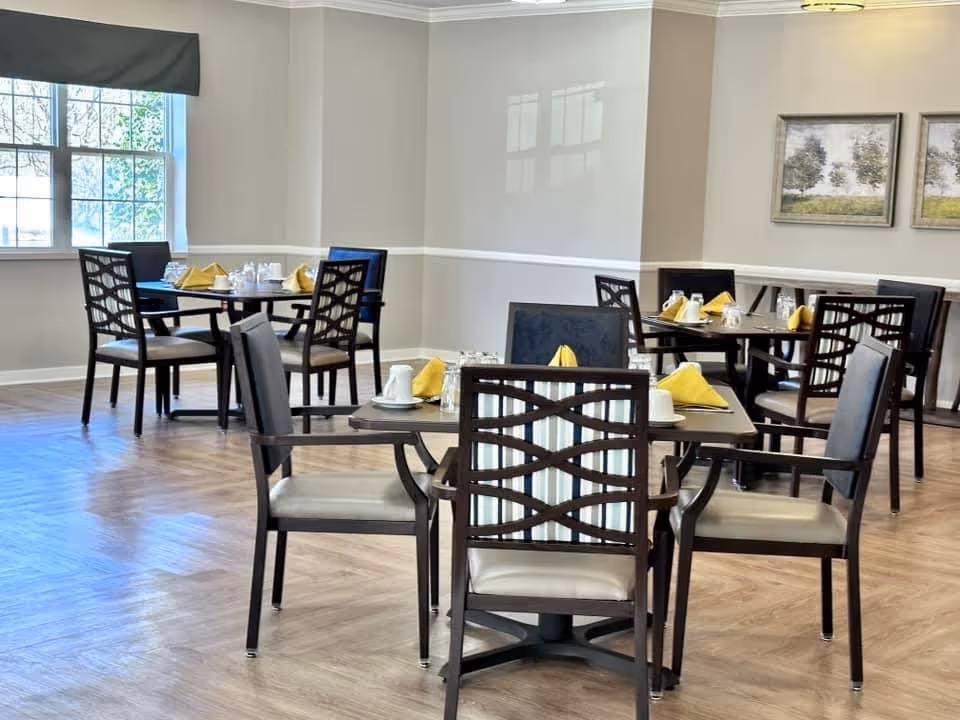 Bright dining room with multiple set tables and chairs, yellow napkins, and large windows.