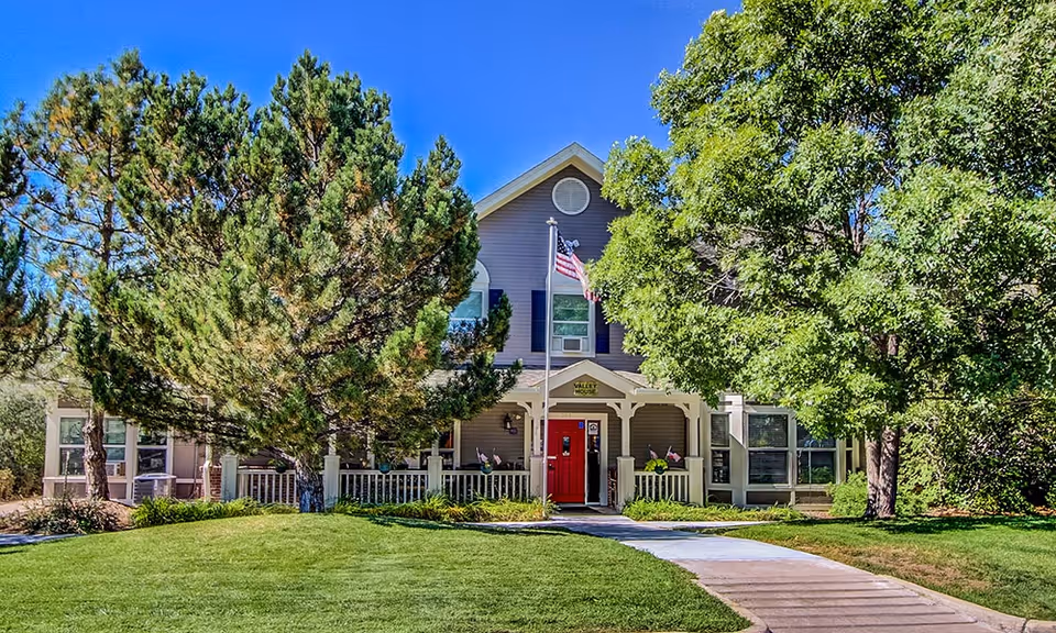 Two-story building with a red front door, covered porch, American flag, and trees on a manicured lawn.