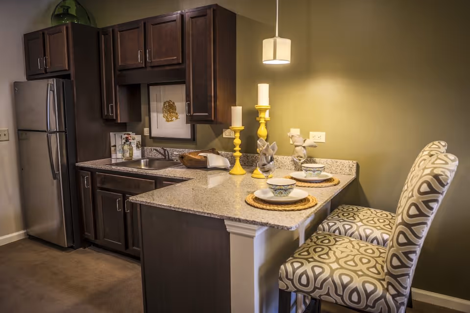 Small kitchen with dark wood cabinets, stainless refrigerator, a granite countertop bar set with two patterned stools and decorative place settings.