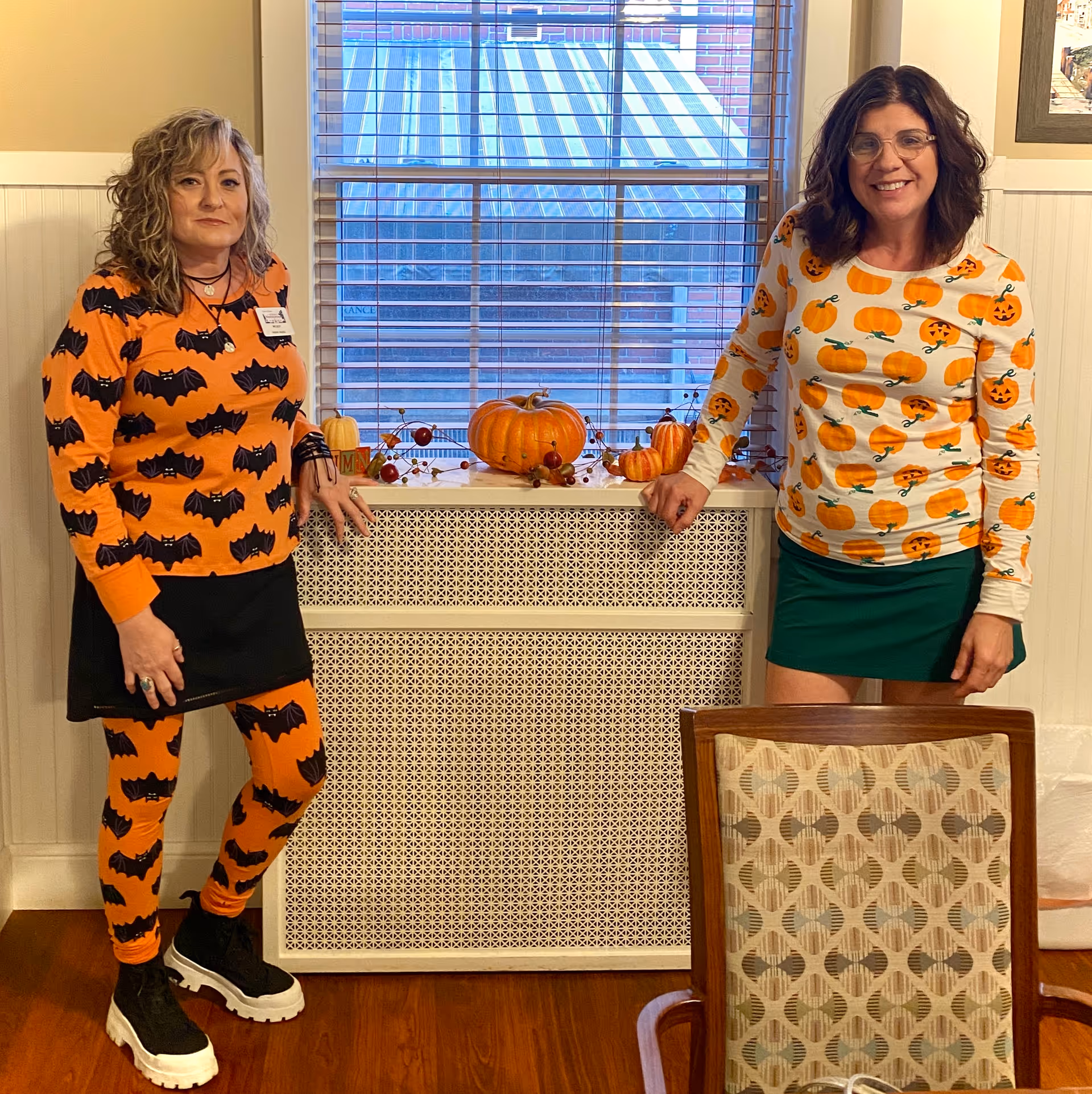 Two women standing indoors in front of a window with blinds. The woman on the left is wearing an orange outfit with black bats, and the woman on the right is wearing a white shirt with orange pumpkins and a green skirt. There are decorative pumpkins and autumn-themed decor on the windowsill behind them. A patterned chair is partially visible in the foreground.