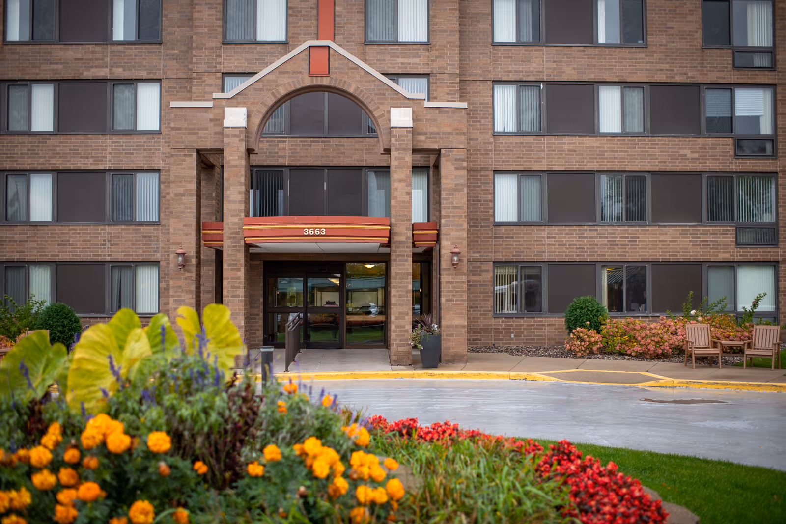 Front exterior view of a multi-story senior living facility with a brick facade, large windows, and an arched entrance canopy displaying the number 3663. In the foreground, there is a colorful flower bed with yellow, orange, red, and purple flowers, along with green foliage. There are also two wooden chairs and some shrubs near the building entrance.