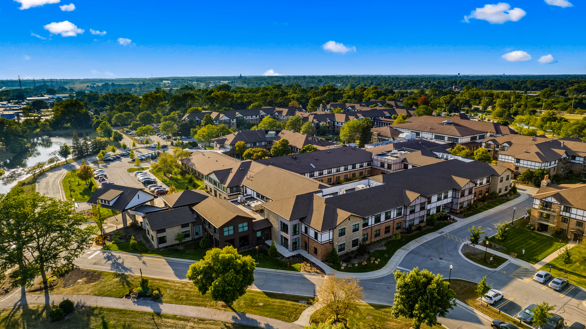 Aerial view of Covenant Living at Windsor Park senior living facility showing multiple buildings with brown roofs surrounded by trees, parking lots with cars, and green landscaped areas under a blue sky with scattered clouds.