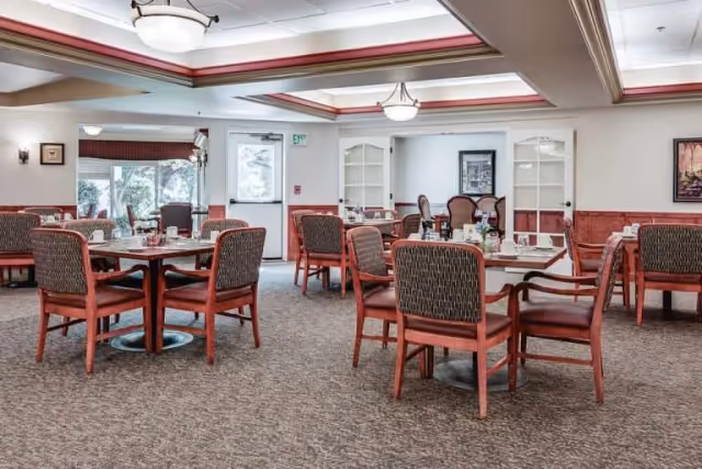 Dining room in a senior living facility with round tables, red chairs, and warm lighting.