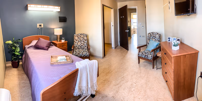 A well-lit bedroom in a senior living facility featuring a single bed with a purple bedspread and pillows, a wooden nightstand with a lamp and tissue box, two patterned armchairs, a wooden dresser with decorative items, and a wall-mounted TV. The room has light-colored walls and flooring, with an open doorway leading to another area.