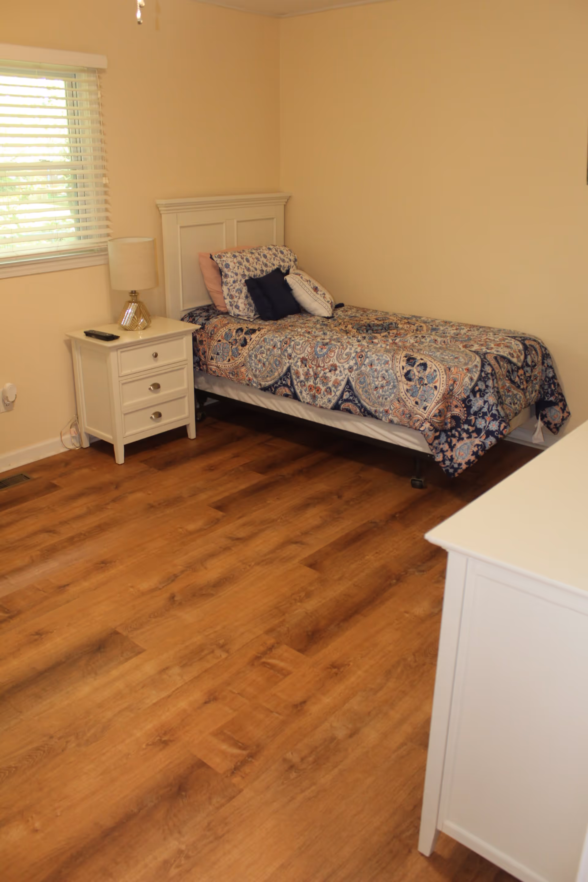 A small bedroom with a single bed covered in a patterned blue, white, and orange bedspread. The bed has multiple pillows and is positioned against a beige wall. Next to the bed is a white nightstand with three drawers, a lamp, and a remote control on top. A window with white blinds is on the left wall, and the floor is wooden.