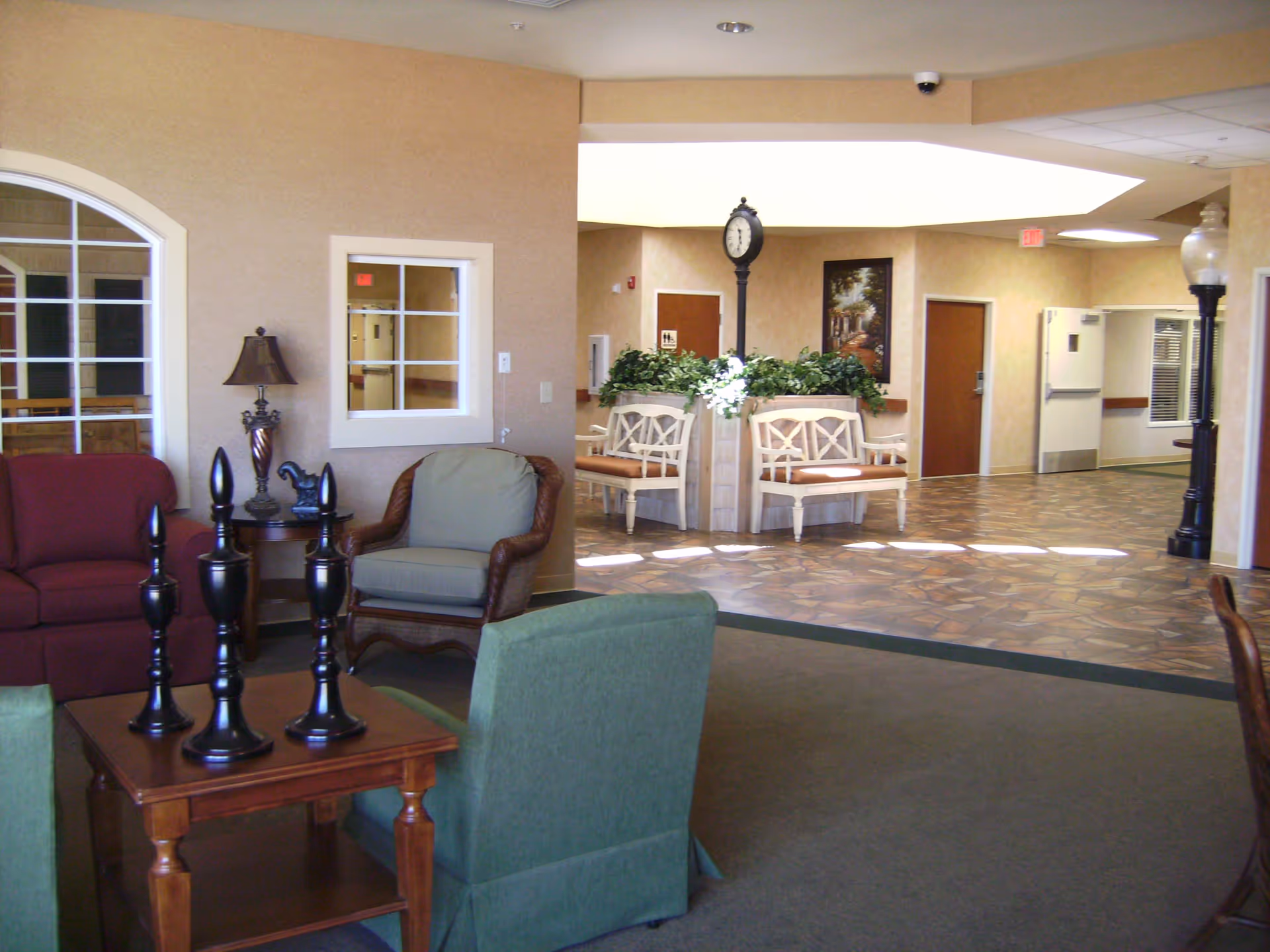 A cozy indoor common area with comfortable chairs, a small wooden table with decorative black finials, a lamp, and a horse figurine. In the background, there are white benches with plants and a tall clock, along with doors and a tiled floor hallway.