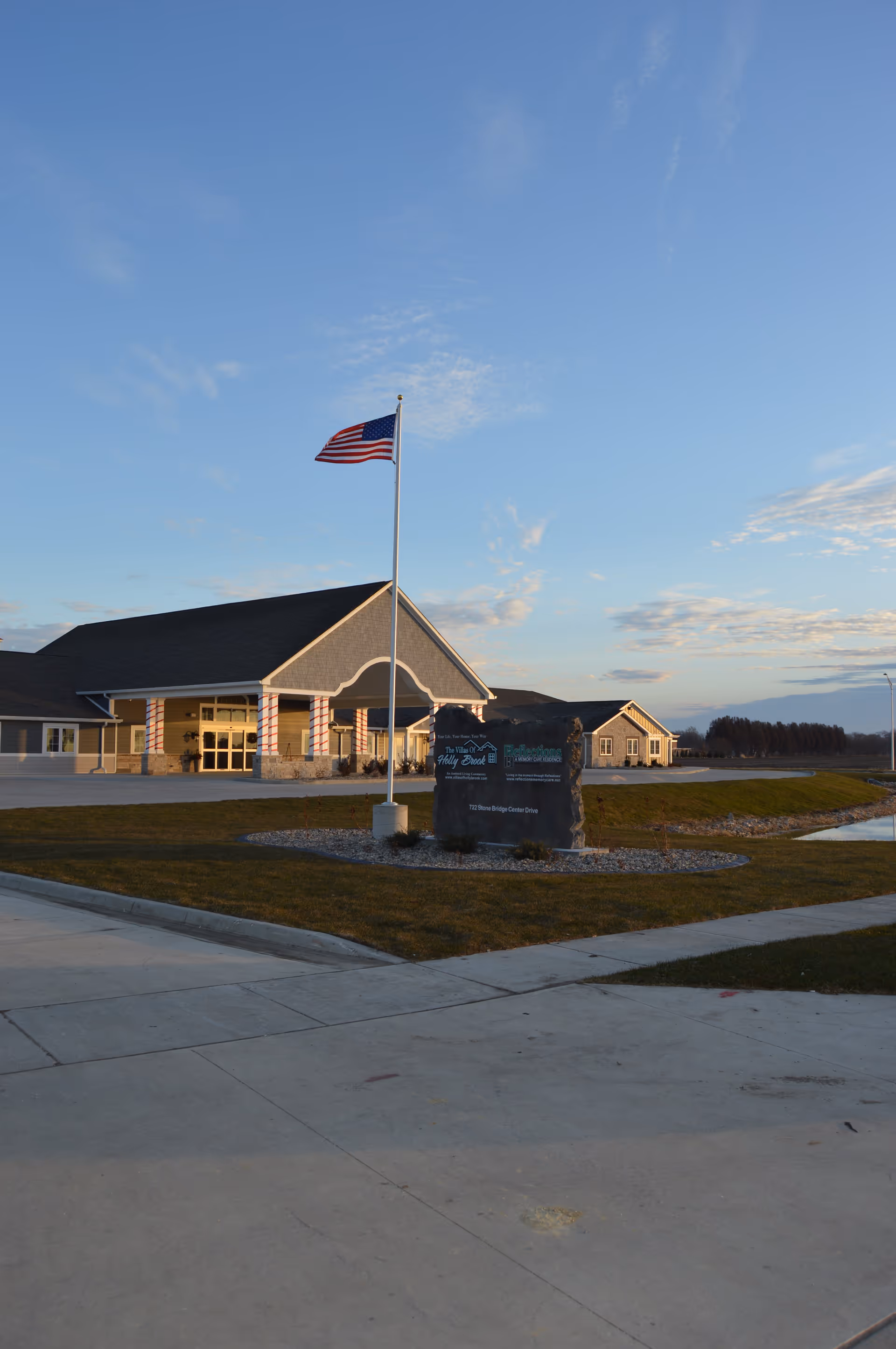 Exterior view of Villas of Holly Brook Assisted Living & Memory Care building in Pekin, IL, with an American flag on a flagpole in front and a stone sign displaying the facility's name and address. The sky is clear with some clouds and the surrounding area includes a paved driveway and landscaped grass.