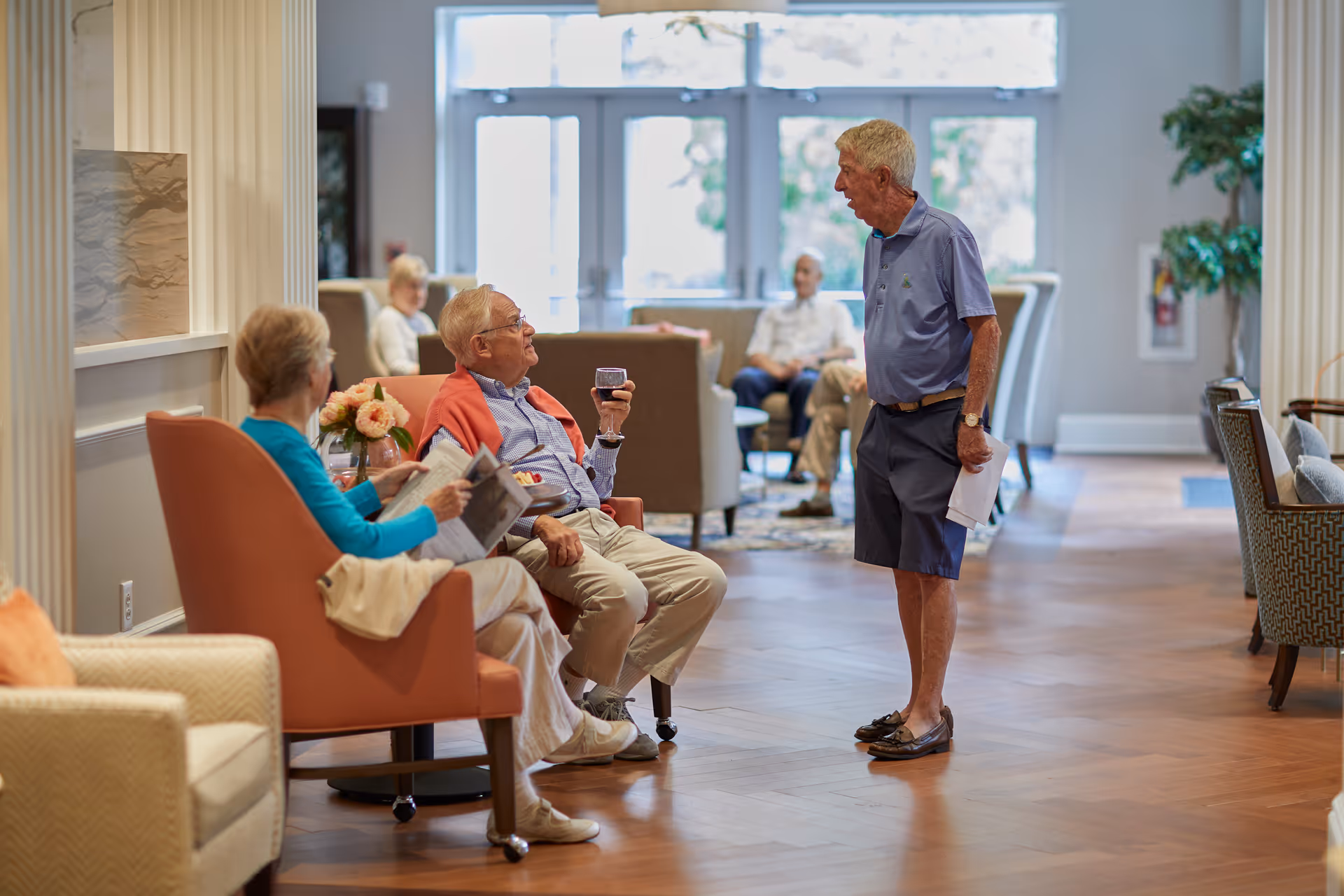 A group of elderly people socializing in a spacious, well-lit common area with comfortable chairs and large windows. One man is standing and talking to a seated man holding a glass of wine, while a woman sits nearby reading a newspaper. Other people are seated in the background, enjoying the relaxed atmosphere.