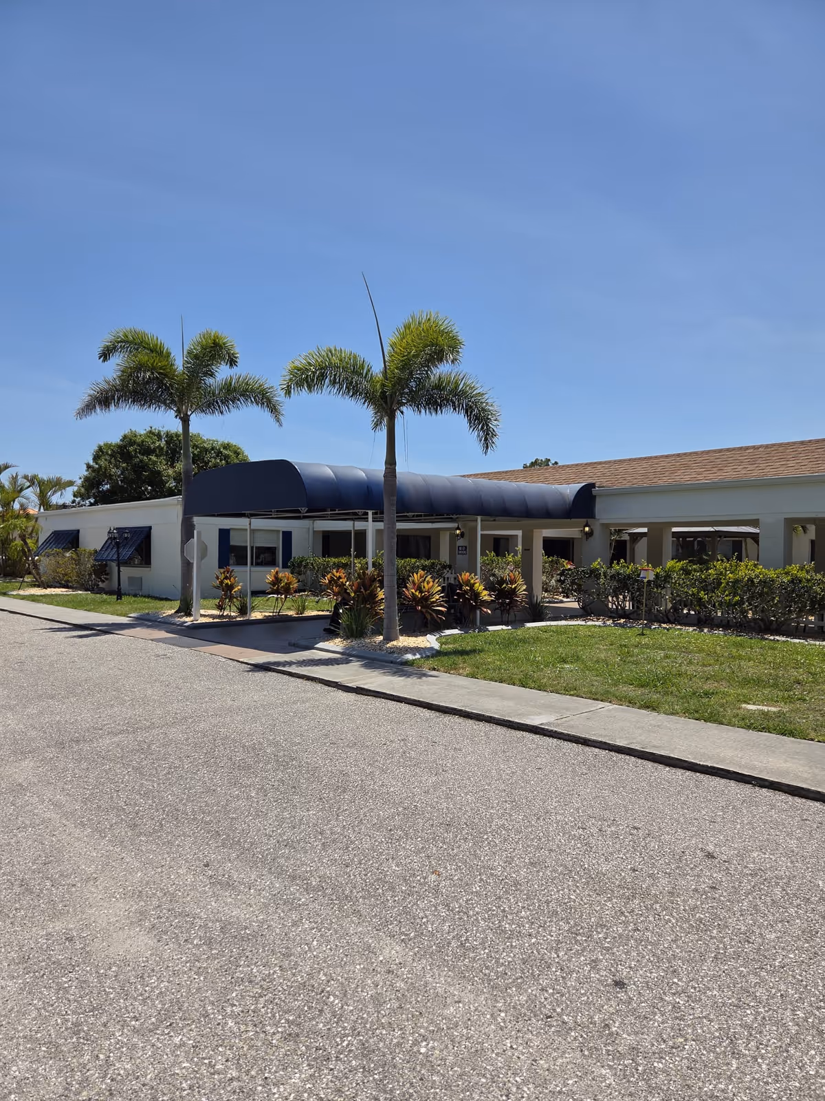 Exterior view of a single-story building with a blue awning over the entrance, surrounded by palm trees and landscaping under a clear blue sky.