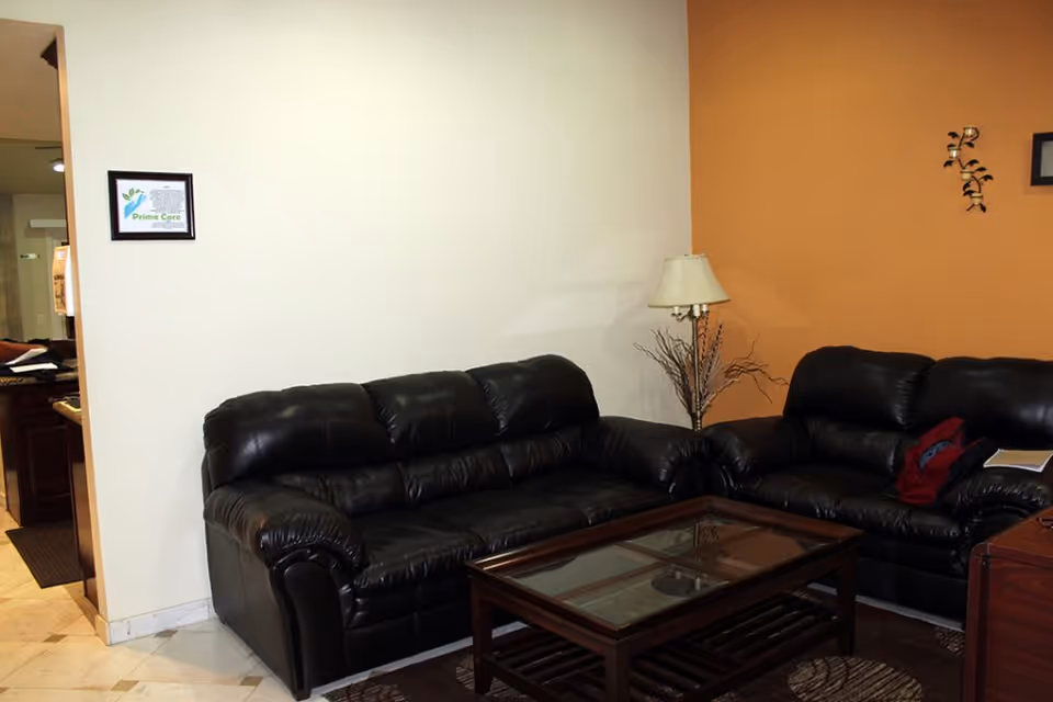 A living room area with two black leather sofas arranged around a glass-top wooden coffee table. There is a floor lamp with a beige shade and decorative dried branches in the corner. The walls are painted white and orange, and a framed certificate or artwork is hung on the white wall. The floor is tiled with a patterned rug under the coffee table.