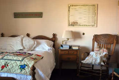A cozy bedroom featuring a wooden bed with white pillows and a colorful quilt. Next to the bed is a wooden nightstand with a lamp and some books. On the wall above the nightstand is a framed family tree chart and a sign that reads 'LOVE IS SPOKEN HERE'. To the right is a carved wooden rocking chair holding two vintage dolls dressed in white.
