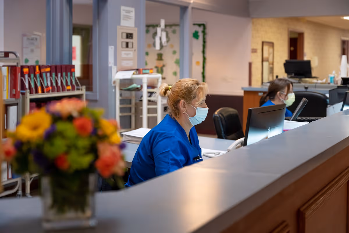 Two healthcare workers wearing blue scrubs and face masks are seated at a reception desk working on computers in a care facility. A colorful bouquet of flowers is visible in the foreground on the desk. The background shows office supplies and a hallway.