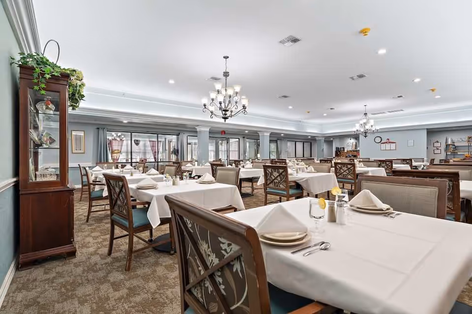 A spacious dining room with tables covered in white tablecloths set with plates and napkins, surrounded by chairs, chandeliers, and a display cabinet.
