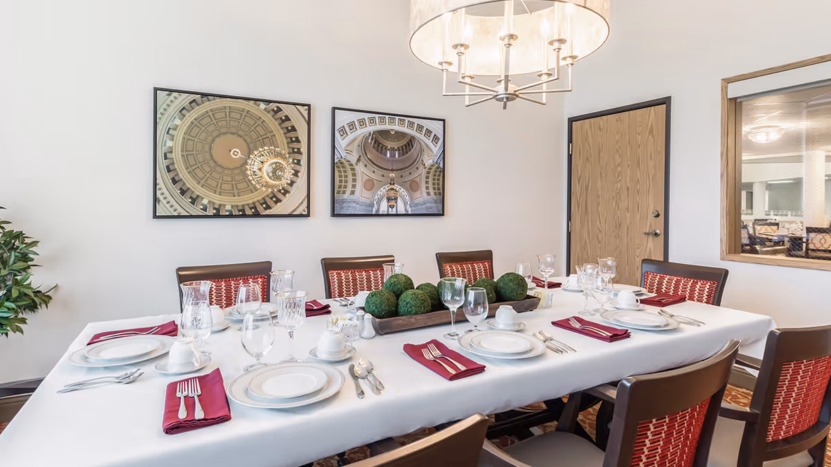 A dining room with a long rectangular table set for eight people. The table is covered with a white tablecloth and has white plates, cups, silverware, wine glasses, and burgundy napkins at each place setting. A centerpiece with green decorative spheres is in the middle of the table. The room has a modern chandelier hanging above the table, two framed pictures of architectural domes on the wall, a wooden door, and a window looking into another room.