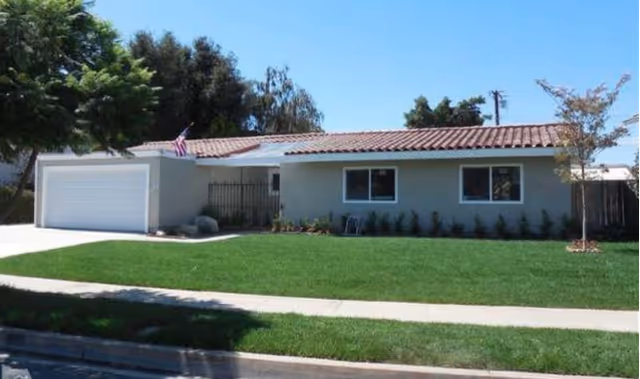 Single-story residential building with a tiled roof, two windows, a garage door, and a well-maintained green lawn in front under a clear blue sky.